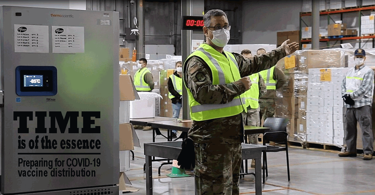 Soldier in uniform, yellow vest and mask, addresses others in warehouse.