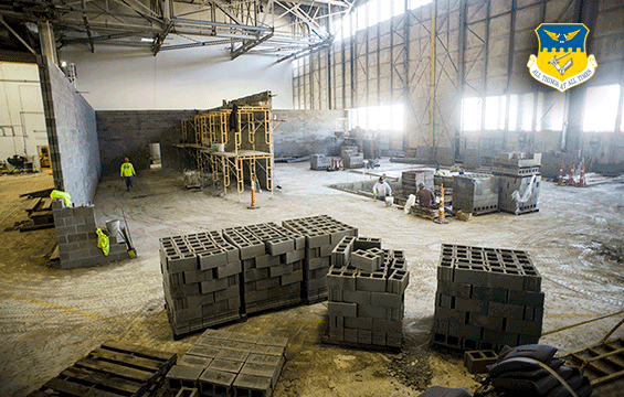Construction site of simulator inside large empty building with pallets of concrete block all around.  Men working with concrete block.