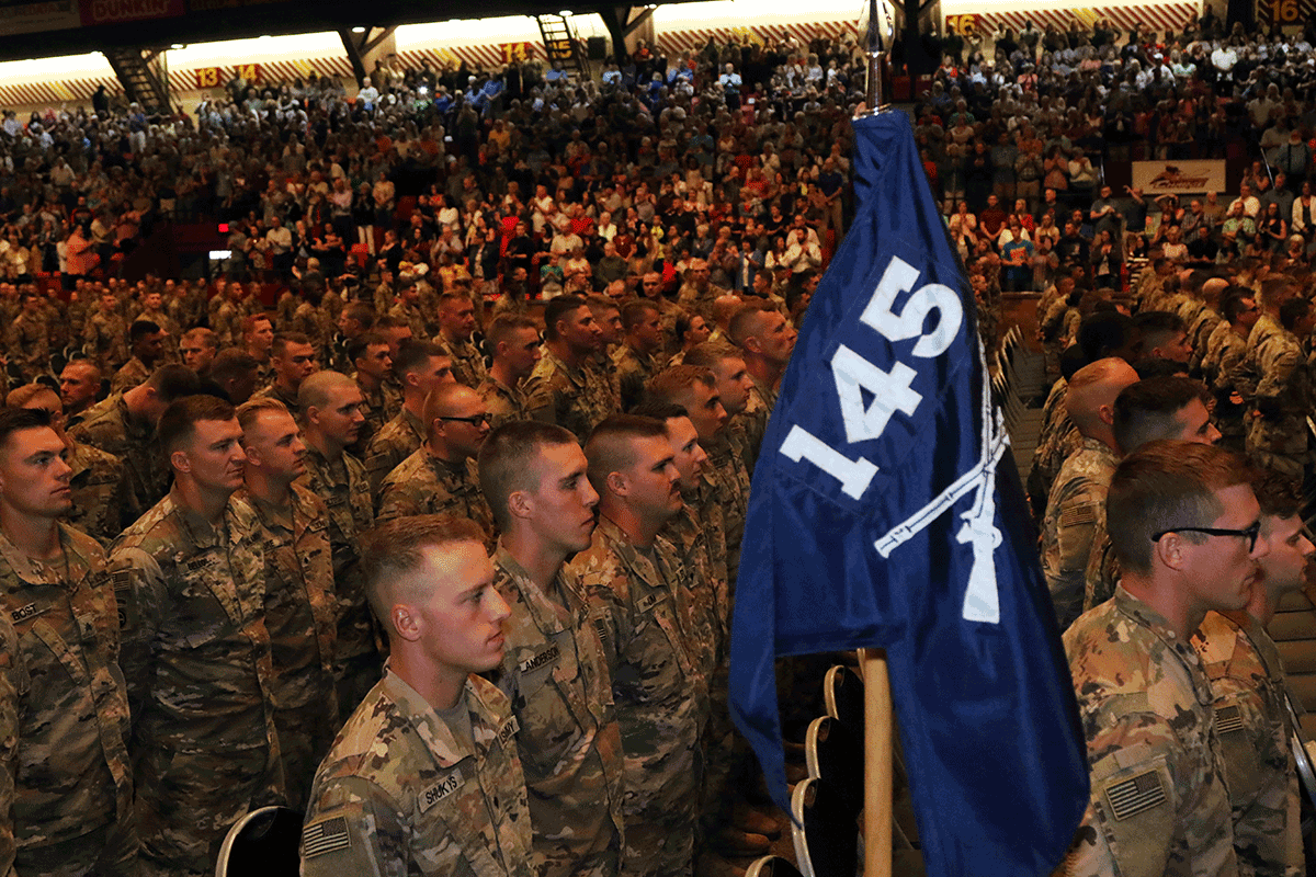 Soldiers stand at attention in auditorium carrying 145th flag.