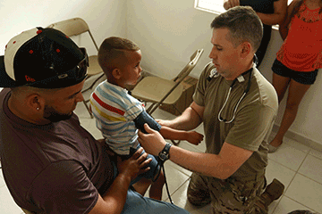 Staff Sgt. Matthew Crabtree, a medic with the 285th Medical Company (Area Support) and a registered emergency room nurse, takes the vital signs of a child sitting on father's lap.