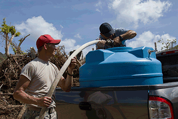 Airman supports hose to local man's blue water container in back of his truck.