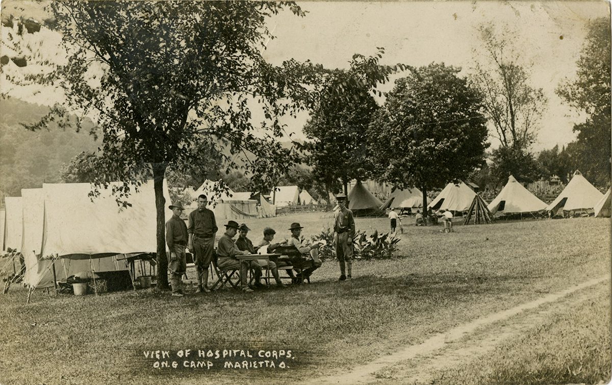 cepia-tone photo of tents under trees with Soldiers gathered around a table