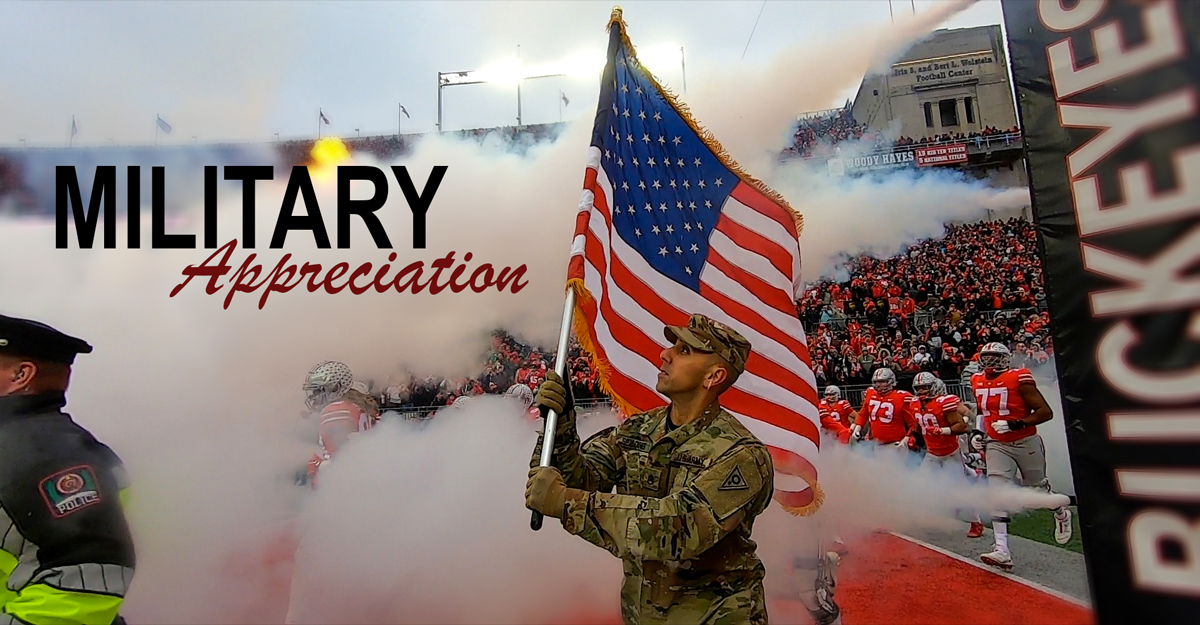 Male Soldier holds up American flag above rising smoke as he leads Ohio State football players onto field.