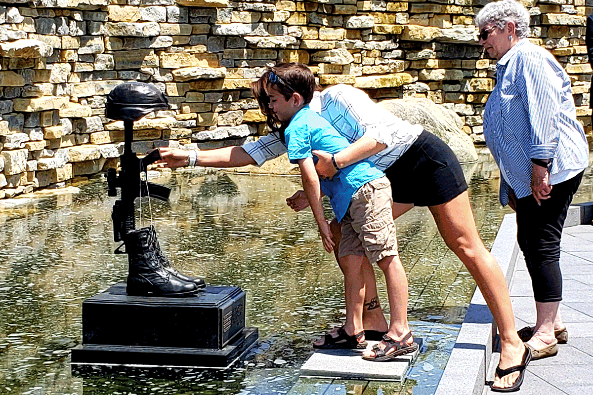 Mother holds son as he reaches to place replica dog tags on the soldier’s cross memorial.