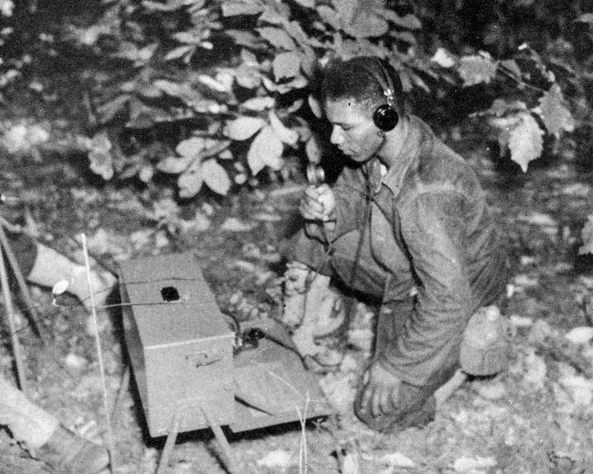 Black soldier kneels to send radio message from field.