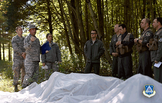 Airmen in treed area stand around parachute on ground as they listen to trainers.