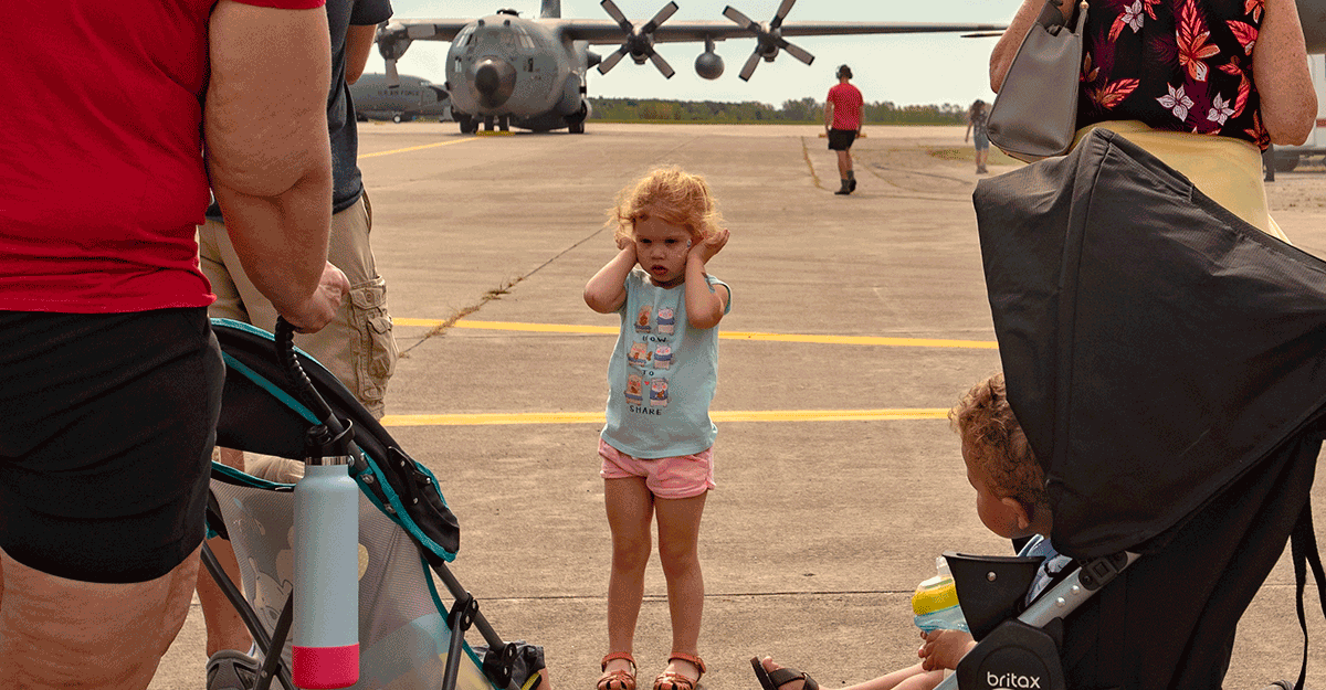 Little girle holding her ears on tarmac with C-130 in background.