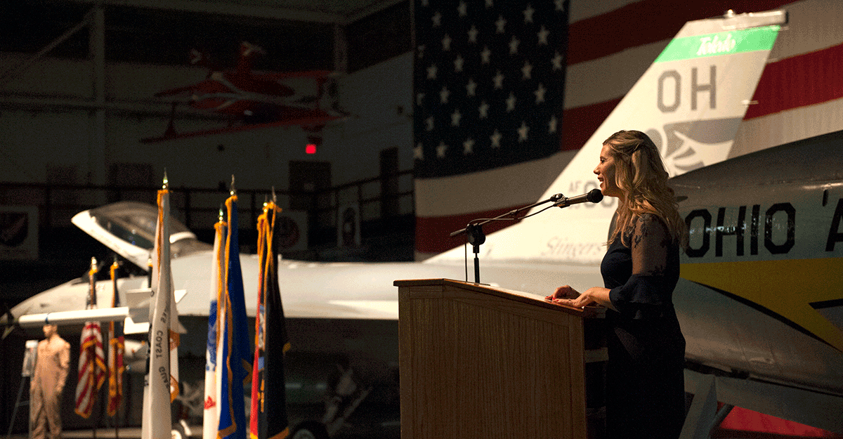 Lorin Sonnenberg-Shaw stands at podium in front of F-16 display.
