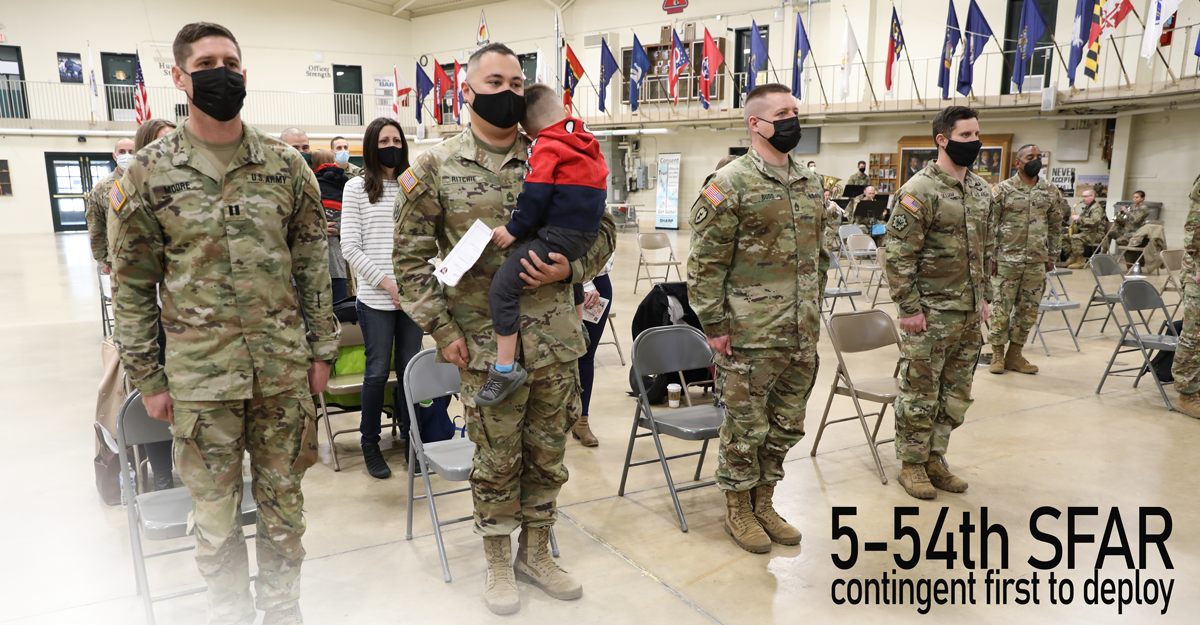 Soldiers with facemasks stand at attention in uniform on drill floor. Male Soldier in front holds child.