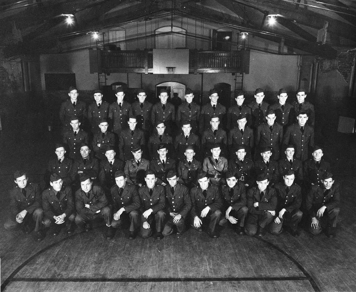 Black and white photo of Soldiers in dress uniform ligned up in rows for group shot inside building.