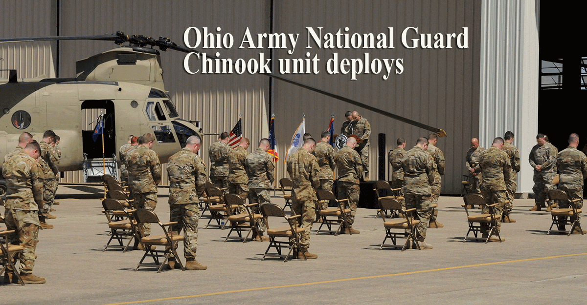 Soldiers stand with heads bowed outside hangar with chinook
