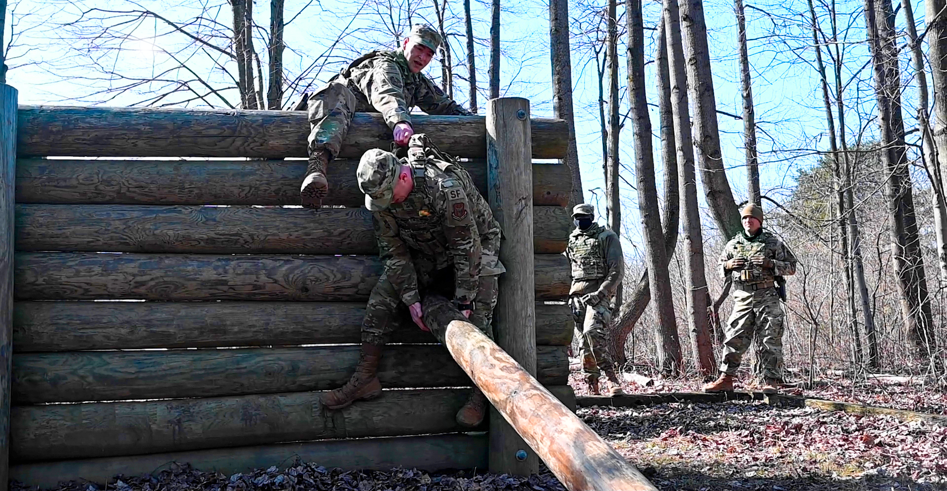 Soldiers navigate a log wall in the woods.