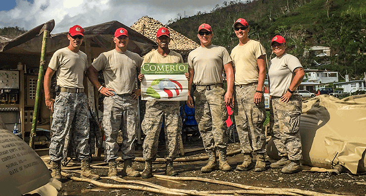 Airmen from the 200th RED HORSE in camo, tshirts and signature red ball caps, stand in front of tent with poster.