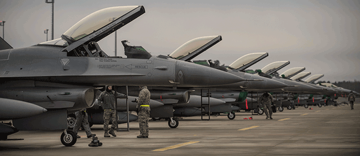 Six F-16s lined up with airmen working around them on ground.