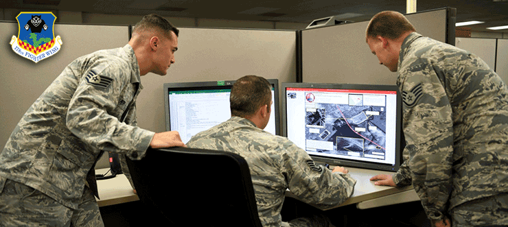 Three airmen galer around desk to look at computer screen.

