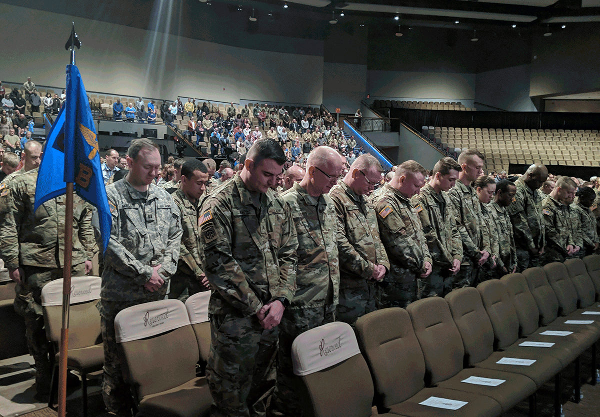 Soldiers in front seats of auditorium standing with heads bowed.