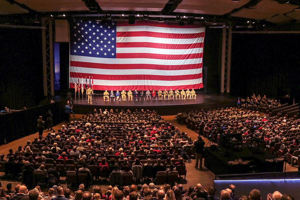 view from back of auditorium of stage with giant American flag as backdrop. Leadership on stage with Soldiers in front rows and guests behind.