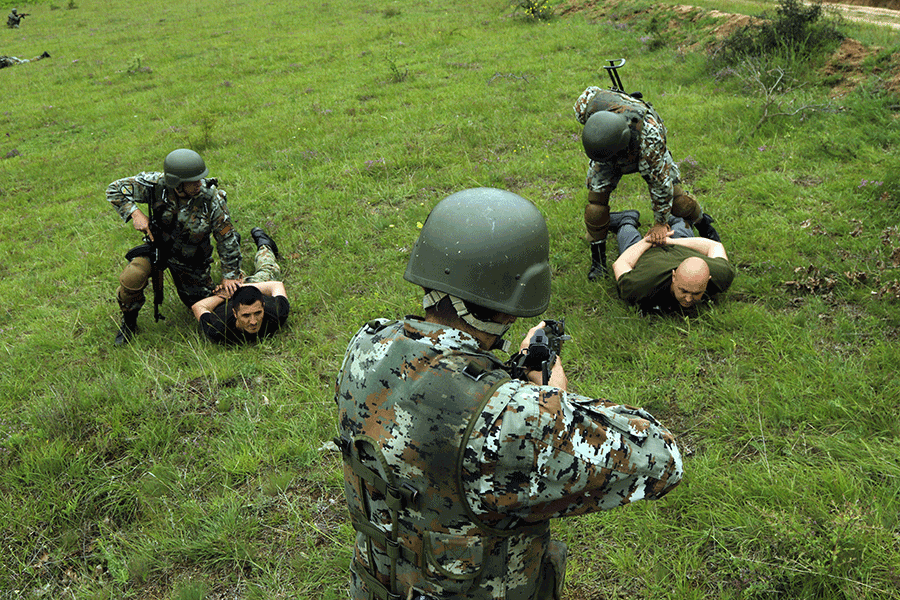 Two soldiers detain men on ground with their hands cuffed behind their back while another soldier secures situation guarding with rifle.