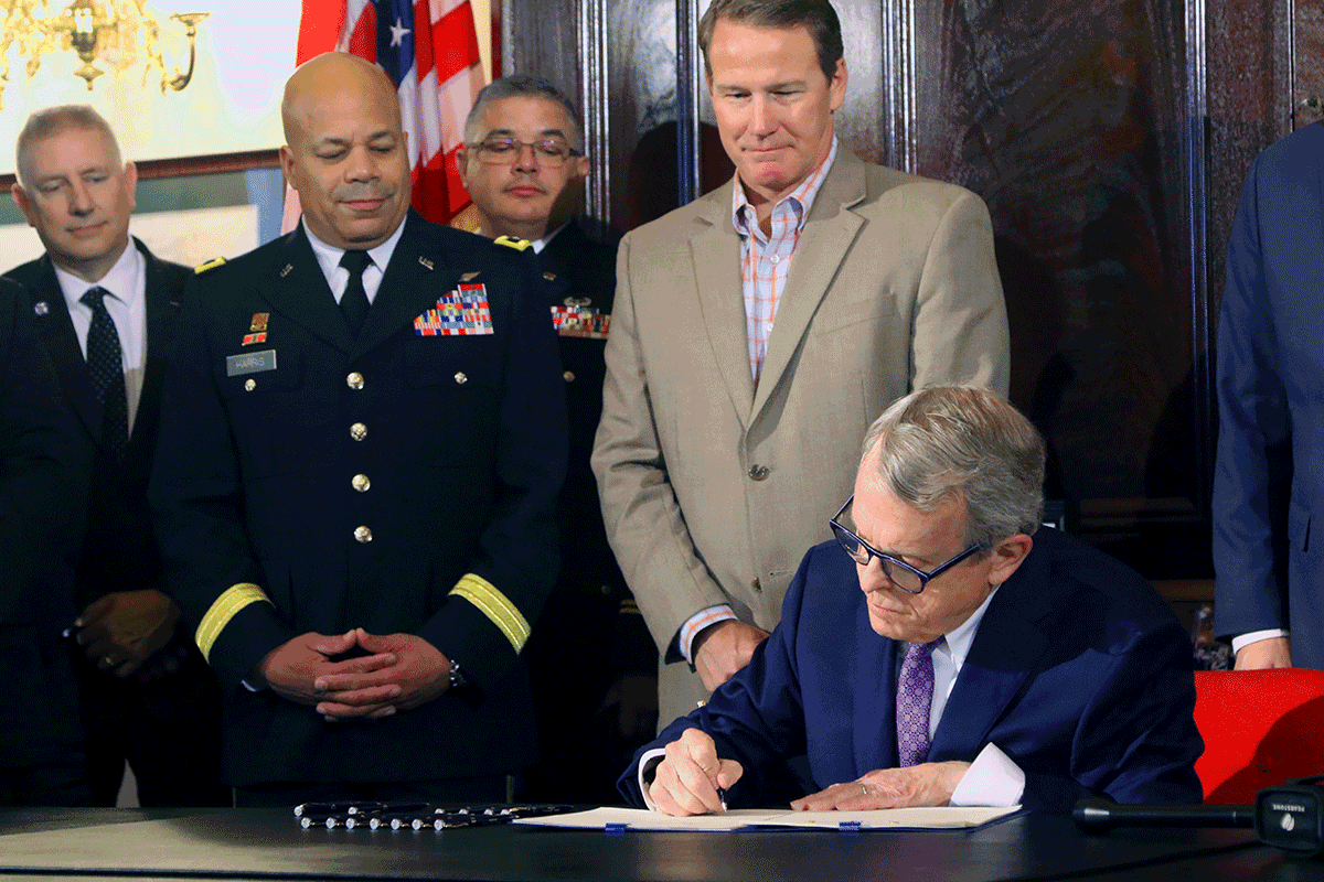 Governor sitting as he signs document while Lt. Gov. Husted, Maj. Gen. John C. Harris, Jr and others gather around table.