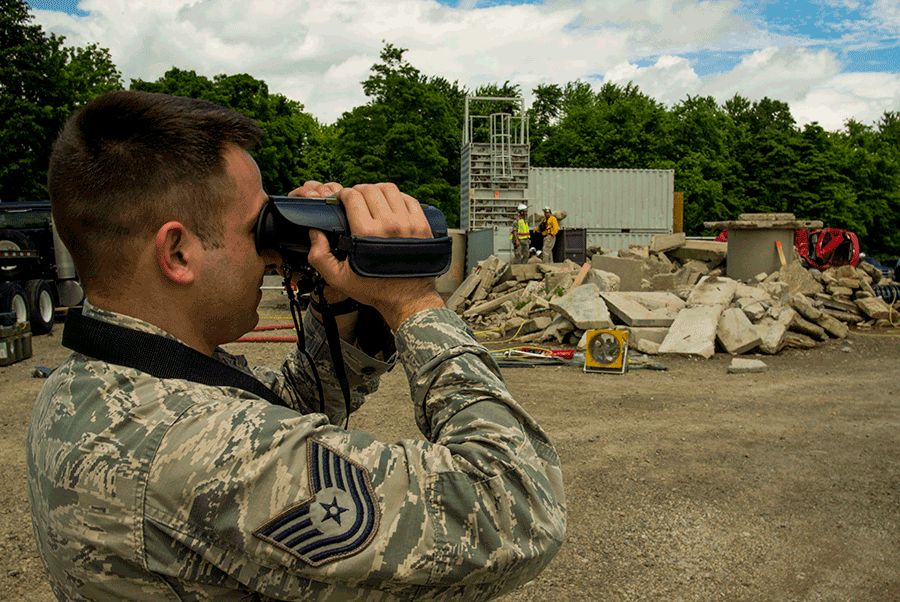 Airman looks through binnoculars at disaster area with rubble in background.