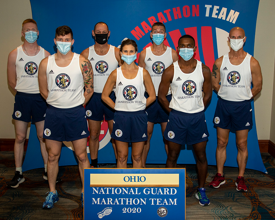 Team in ONG tanks and shorts pose with mask in front of 2020 Marathon Team plaque.