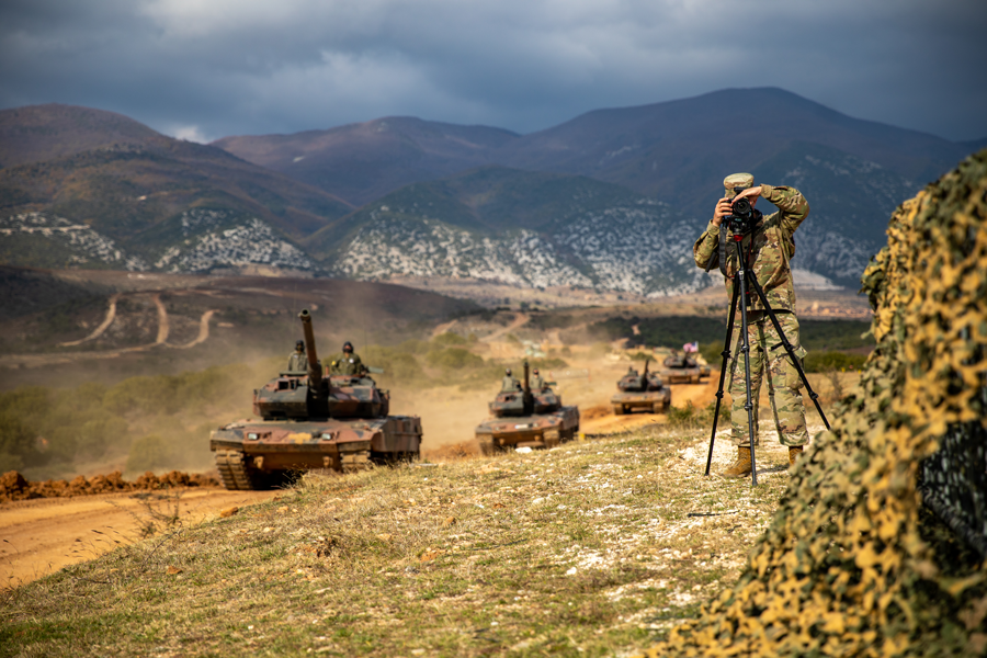Soldier sets up tripod in Greece with tanks caravanning beside and mountains.
