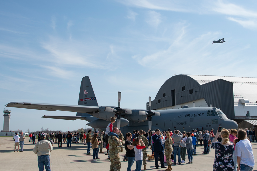 Crowd gathered around a C-130 while another C-130 flies over hangar.