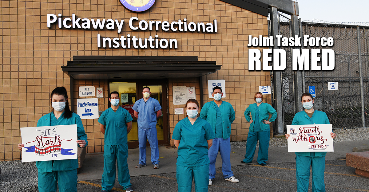 Members of Joint Task Force Red Med pose in front of Pickaway Correctional Institute wearing scrubs, masks and holding signs of support.
