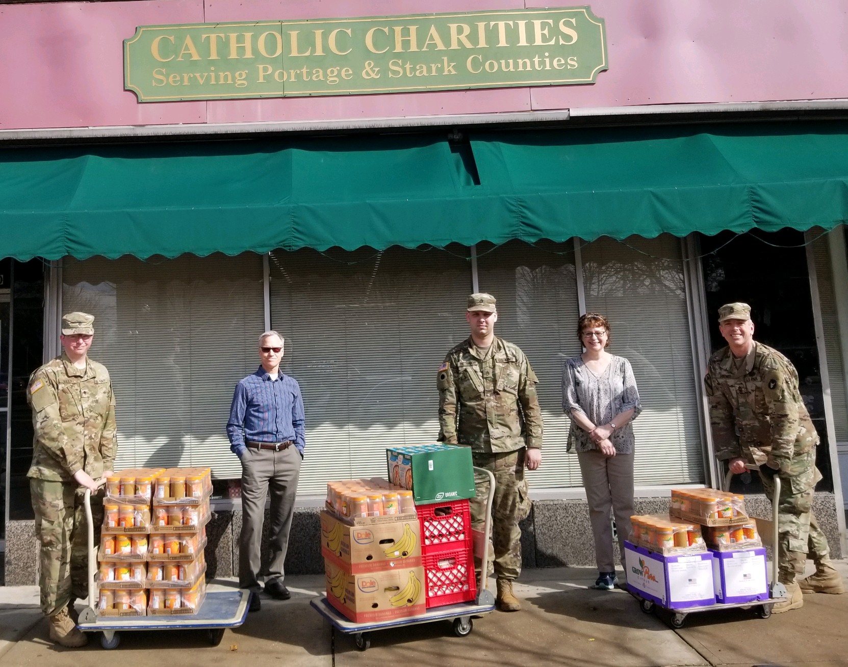 Soliders stand with civilians with pallets of food in front of Catholic Charities pantry.